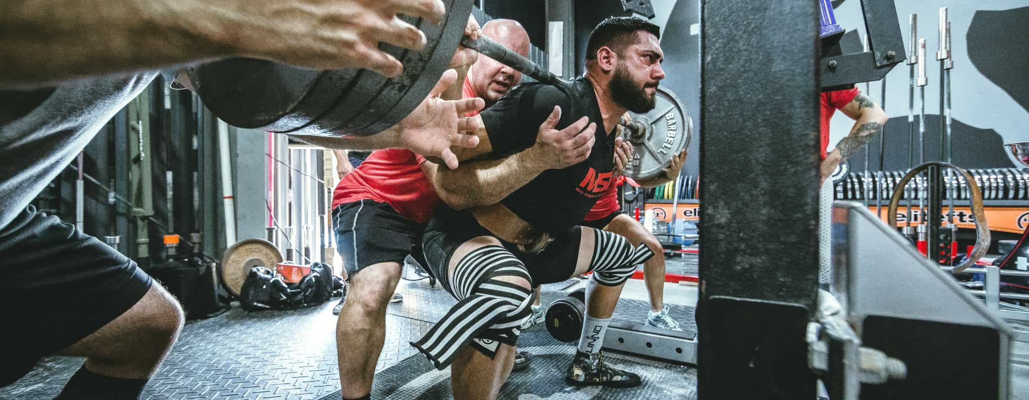 A man doing a heavy squat with a spotter behind him and a spotter at each end of the bar.