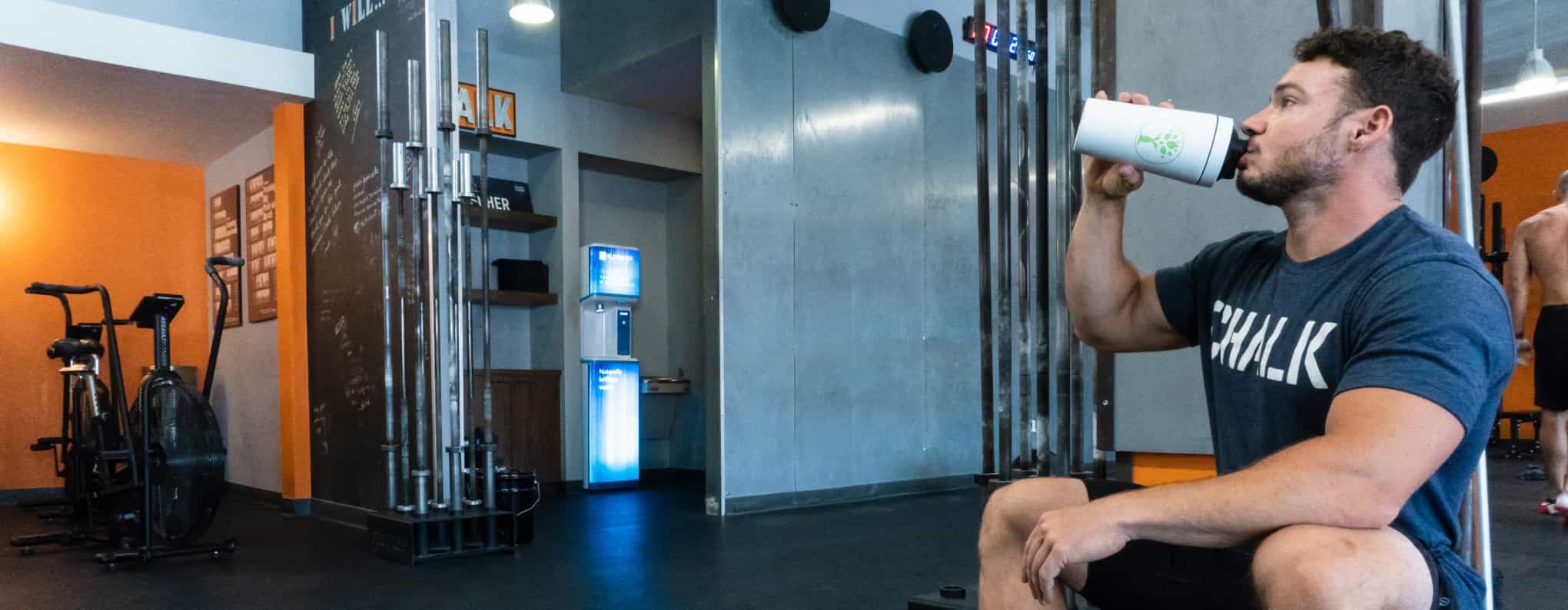 A bearded man sitting on a weights bench drinking from a protein shaker