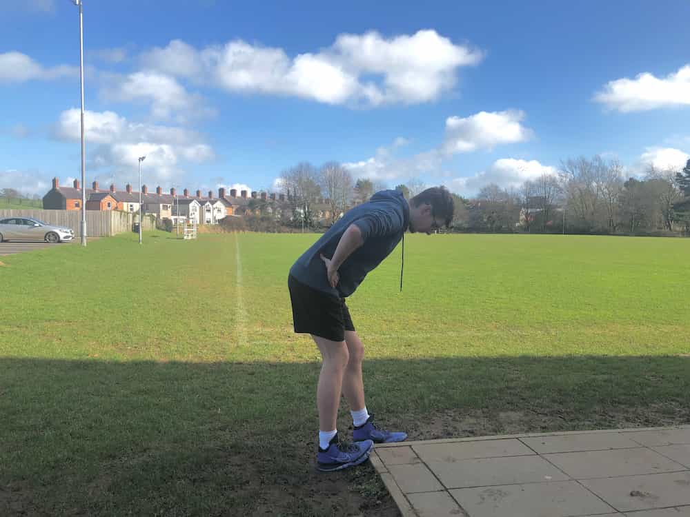 Jordan doing a soleus stretch beside a football pitch on a sunny day. He is wearing a petrol blue hoodie and black shorts.
