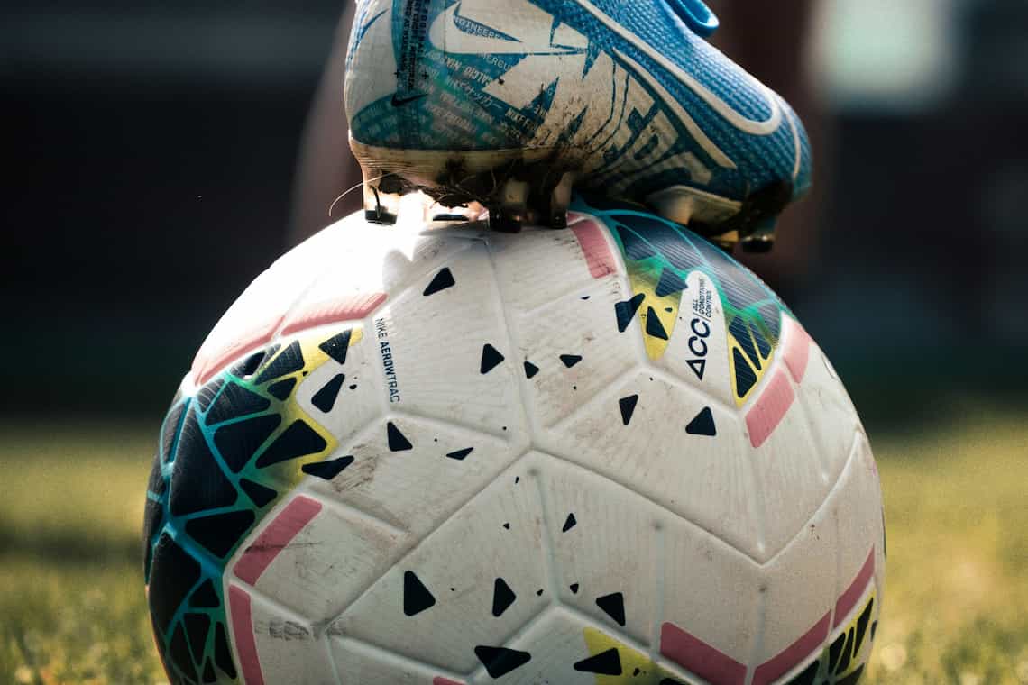 A close up of a ball with the foot of a player resting on top of it. The ball is white with small patches of blue, black and red, and the boot is blue.