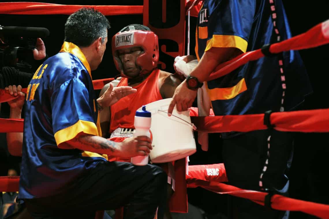 A boxer on the stool between rounds getting advice from his corner. The boxer is in a red headguard, red single and red shorts.