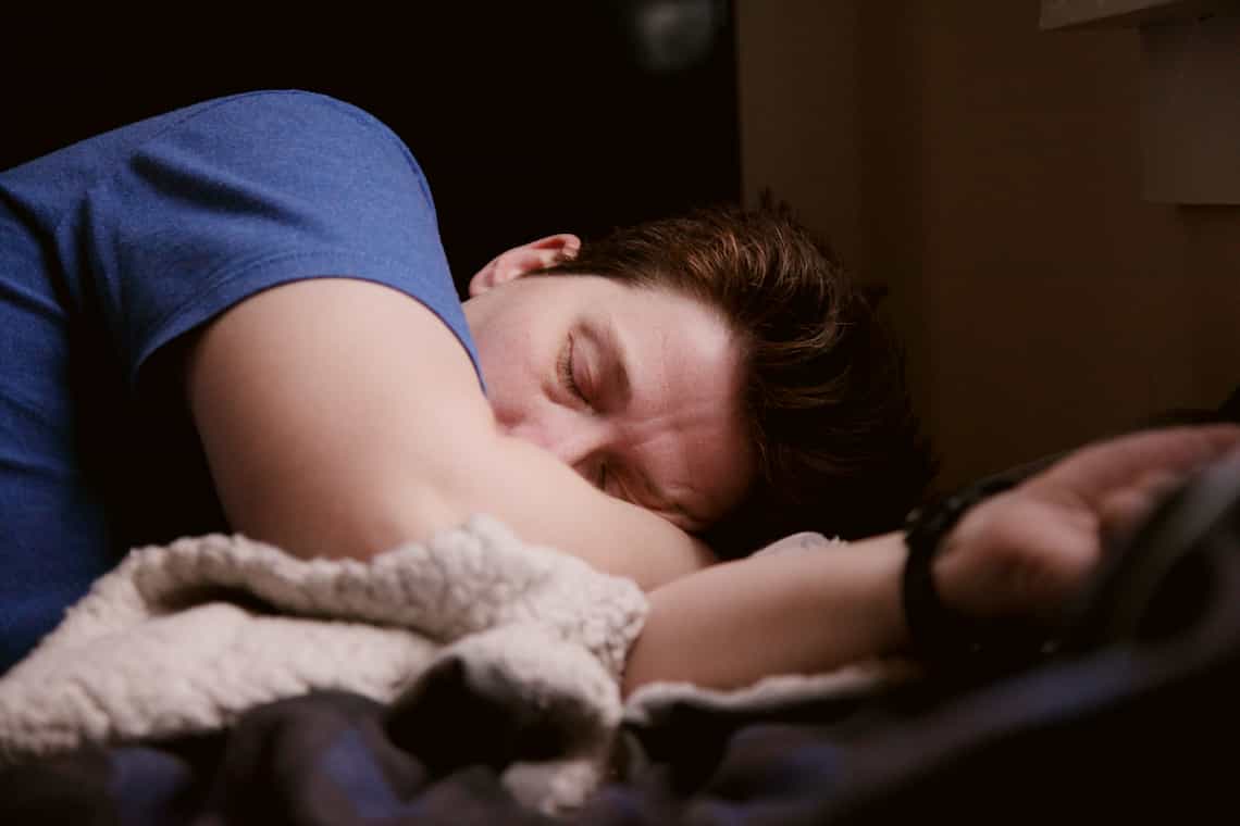 A man in a blue tshirt lying down with his eyes closed and a slight frown.